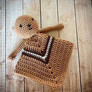 May include: A handmade crochet doll in shades of brown, with a square blanket. The doll has a round head, small black eyes, and outstretched arms. The blanket features a V-neck design with brown and white stripes.