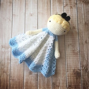 May include: A crocheted baby doll with blonde hair and a black bow. The doll is wearing a blue and white dress and is lying on a wooden surface.