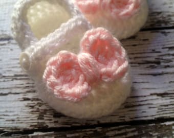 May include: A pair of white crocheted baby booties with pink crocheted bows. The booties are on a whitewashed wooden surface.
