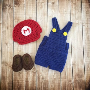 Pu&ograve; includere: Un completo per neonato all'uncinetto a tema Super Mario. Il completo comprende un cappello rosso con una 'M' bianca, una salopette blu con bottoni gialli e scarpine marroni.