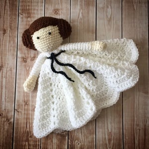 May include: White crocheted baby blanket with a crocheted doll wearing a white dress and black sash. The doll has brown hair and brown eyes.