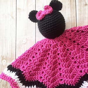 May include: A pink and black crocheted baby blanket with a black crocheted mouse head attached. The mouse head has a pink bow.