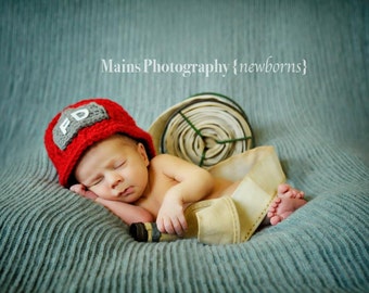May include: A newborn baby wearing a red crocheted firefighter hat with the letters "FD" on it, sleeping on a blue blanket with a fire hose prop.