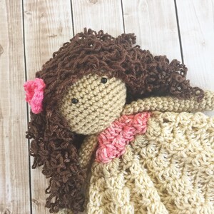Pode incluir: Uma boneca de croch&ecirc; com cabelo encaracolado castanho, uma flor rosa e um vestido bege com um detalhe rosa.