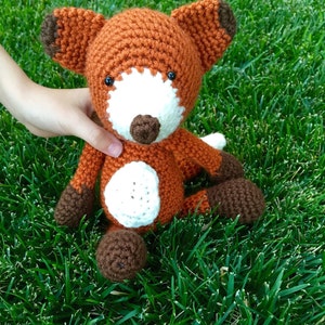 May include: A crocheted fox plush toy with brown, orange, and white fur. The fox is sitting on a bed of green grass.