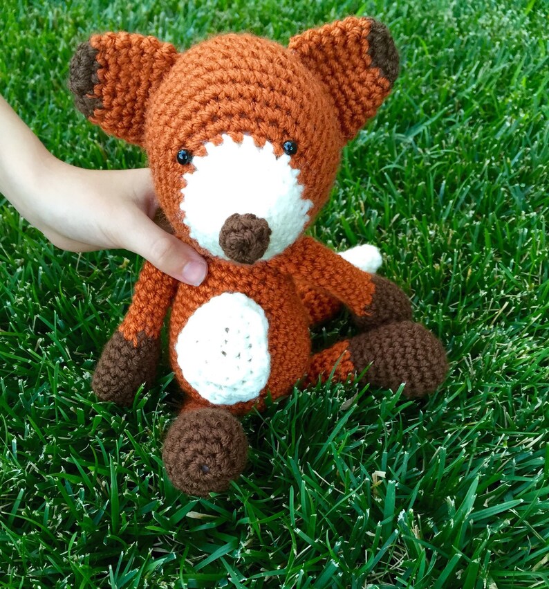 May include: A crocheted fox plush toy with brown, orange, and white fur. The fox is sitting on a bed of green grass.