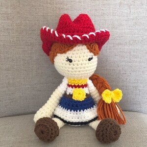 May include: Crocheted doll with brown hair in a braid with a yellow bow, wearing a white, blue, and red dress with a yellow star, and a red cowboy hat with white trim.