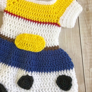 以下が含まれることがあります： 白、黄、青、赤、茶色、黒のクロシェ編みのベビーワンピース。ワンピースは、前面に黄色の楕円形がある漫画のキャラクターのようにデザインされています。