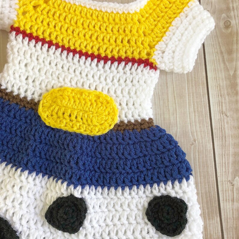 以下が含まれることがあります： 白、黄、青、赤、茶色、黒のクロシェ編みのベビーワンピース。ワンピースは、前面に黄色の楕円形がある漫画のキャラクターのようにデザインされています。