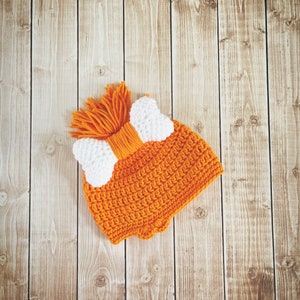May include: Orange crocheted hat with a white bow on a wood background.