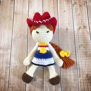 May include: Crocheted doll with red hair, wearing a red and white cowboy hat, a blue and white dress, and a yellow sheriff's star. The doll has brown eyes and a brown braid with a yellow bow.