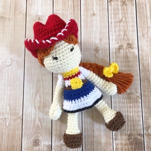 May include: Crocheted doll wearing a red and white cowboy hat, a blue and white dress, and a yellow sheriff's star. The doll has brown hair and black eyes.