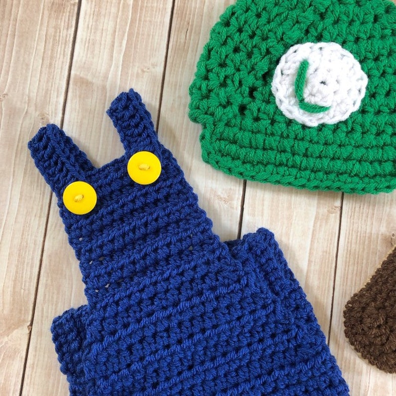 May include: Blue crocheted overalls with yellow buttons and a green crocheted hat with a white L on it.