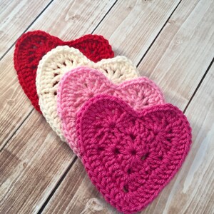 Op de afbeelding: Vier gehaakte harten in tinten van rood, roze en wit. De harten zijn op elkaar gelegd, met de grootste hart onderaan en de kleinste hart bovenaan.