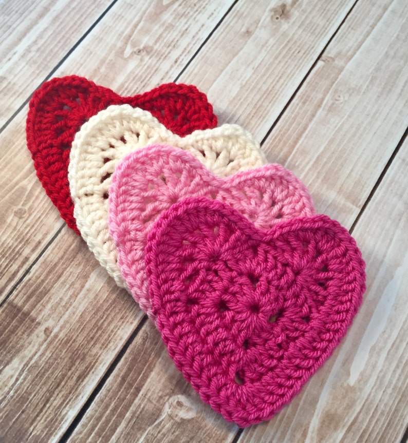 Op de afbeelding: Vier gehaakte harten in tinten van rood, roze en wit. De harten zijn op elkaar gelegd, met de grootste hart onderaan en de kleinste hart bovenaan.