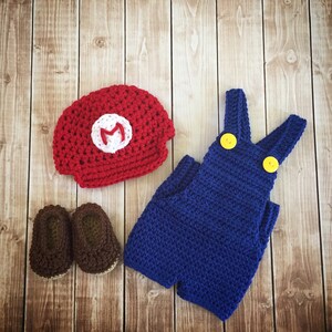 Pu&ograve; includere: Un completo per neonato all'uncinetto ispirato al personaggio del videogioco Mario. Il completo comprende un cappello rosso con una 'M' bianca, una salopette blu con bottoni gialli e scarpe marron.