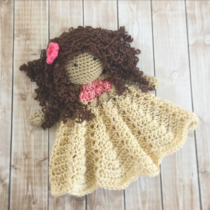 Pode incluir: Uma boneca de croch&ecirc; com cabelo castanho encaracolado, uma flor rosa e um vestido rosa e bege. A boneca est&aacute; usando um vestido de croch&ecirc; bege e est&aacute; deitada em uma superf&iacute;cie de madeira.