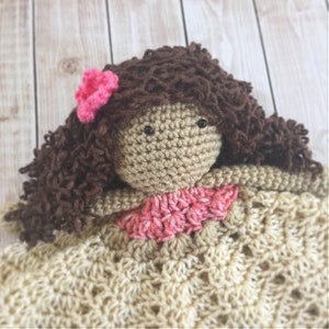 Pode incluir: Uma boneca de croch&ecirc; com cabelo castanho encaracolado, uma flor rosa e um cobertor de croch&ecirc; bege. A boneca tem olhos castanhos e um vestido de croch&ecirc; rosa e bege.