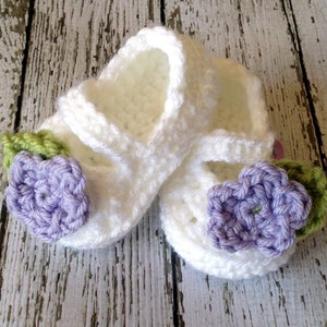 May include: A pair of white crocheted baby shoes with purple and green flower embellishments.