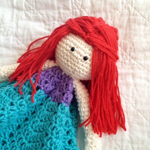 May include: A crocheted doll with long red hair wearing a turquoise and purple dress. The doll is lying on a white quilted fabric.