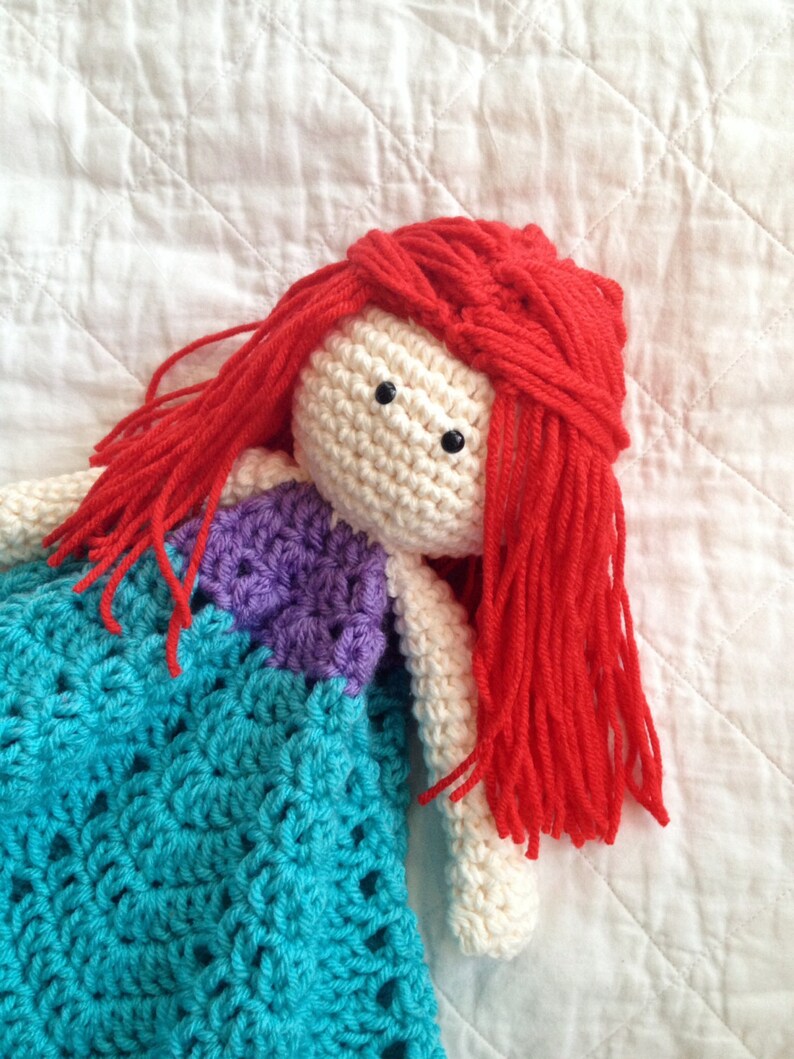 May include: A crocheted doll with long red hair wearing a turquoise and purple dress. The doll is lying on a white quilted fabric.