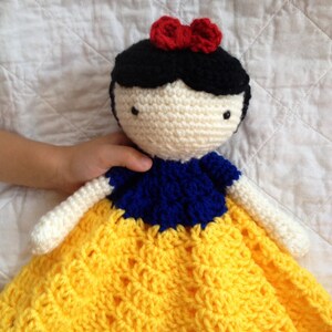 May include: A crocheted Snow White doll with a yellow dress, blue bodice, and red bow. The doll is lying on a white blanket.