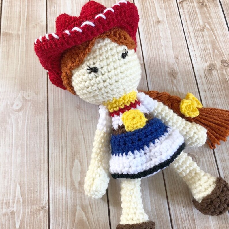 以下が含まれることがあります： 赤、白、青の服を着た、かぎ針編みの人形。人形は茶色の髪、赤いカウボーイハット、黄色の星型の保安官バッジをしています。