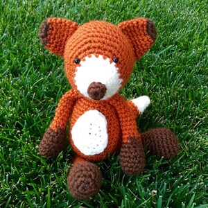 May include: A crocheted fox stuffed animal with orange fur, white chest, and brown accents. The fox is sitting on a bed of green grass.