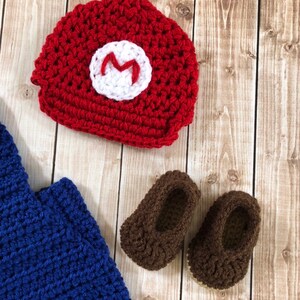 May include: A crocheted baby outfit in red, blue, and brown. The outfit includes a hat with an 'M' on the front, a blue jumpsuit, and brown booties.
