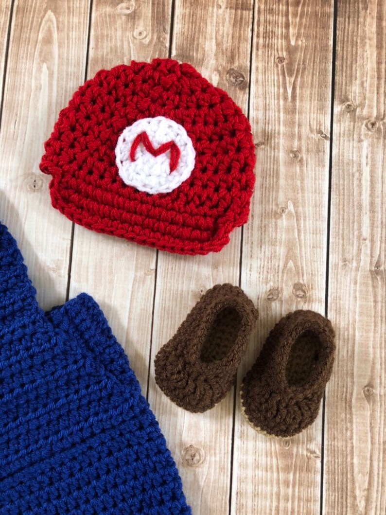 May include: A crocheted baby outfit in red, blue, and brown. The outfit includes a hat with an 'M' on the front, a blue jumpsuit, and brown booties.