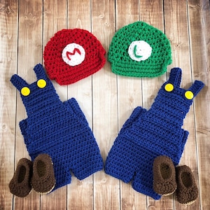 May include: Two blue crocheted overalls with yellow buttons and matching crocheted shoes. One red and one green crocheted hat with white letters 'M' and 'L' on the front.