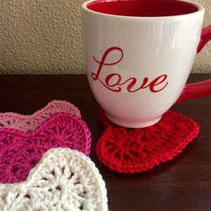 Op de afbeelding: Een witte mok met het woord "Love" in rood op de zijkant, die op een rode gehaakte hartvormige onderzetter staat. Op de achtergrond zijn drie extra gehaakte hartvormige onderzetters te zien, in roze en witte tinten.