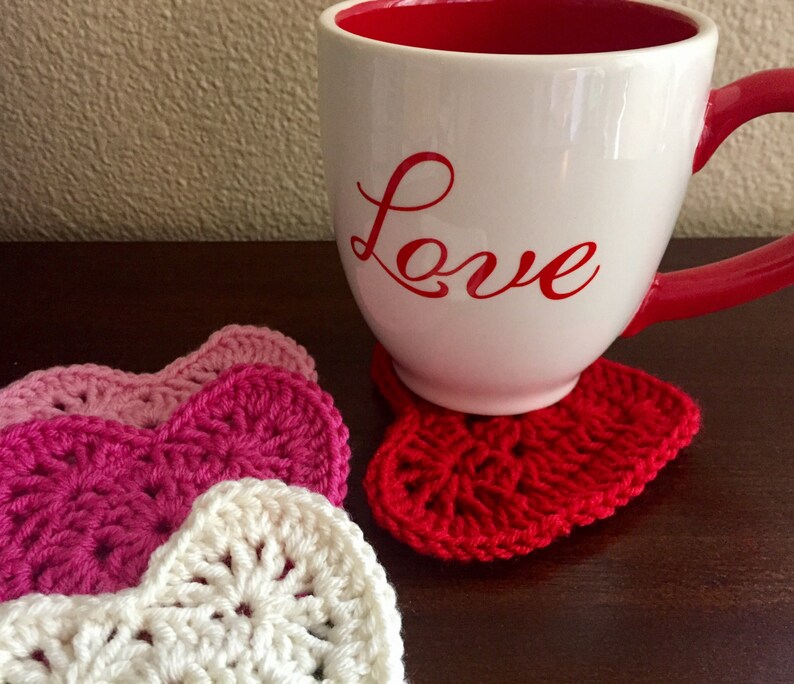 Op de afbeelding: Een witte mok met het woord "Love" in rood op de zijkant, die op een rode gehaakte hartvormige onderzetter staat. Op de achtergrond zijn drie extra gehaakte hartvormige onderzetters te zien, in roze en witte tinten.
