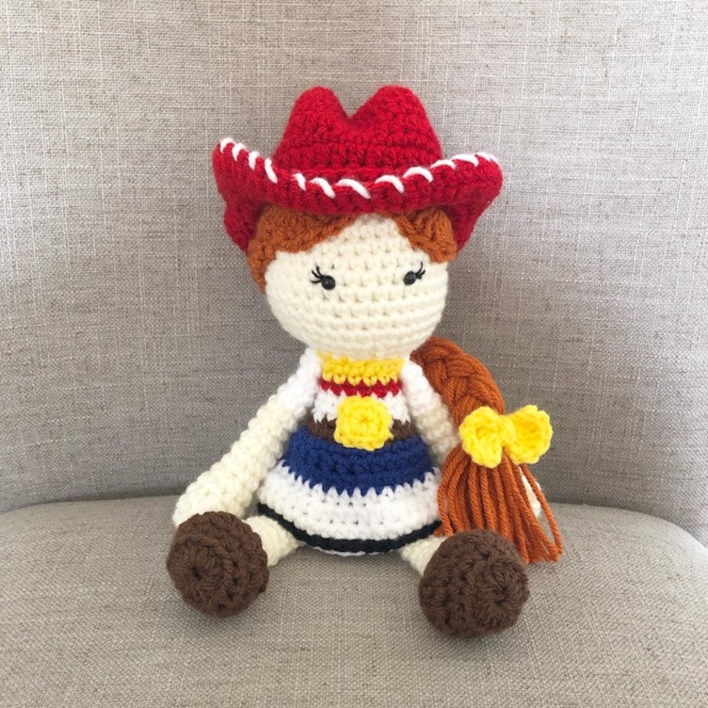 May include: Crocheted doll with brown hair, wearing a red cowboy hat with white trim, a blue and white dress, and a yellow star sheriff's badge. The doll has brown yarn for legs and a yellow bow in its hair.