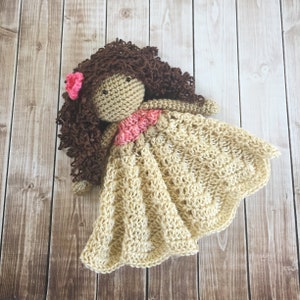 Pode incluir: Uma boneca de croch&ecirc; com cabelo castanho encaracolado, uma flor rosa e um vestido bege. A boneca est&aacute; deitada sobre uma superf&iacute;cie de madeira.