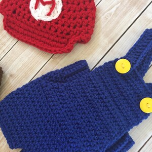 Pu&ograve; includere: Salopette in crochet blu con bottoni gialli e un cappello in crochet rosso con una M bianca.