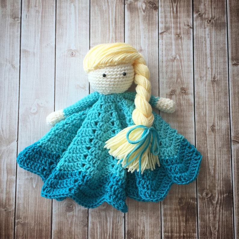 May include: A crocheted baby blanket with a doll attached. The doll has a white face, black eyes, and long blonde hair. The blanket is blue and green with a scalloped edge.