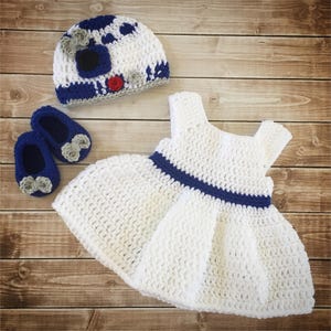 May include: A white crocheted dress with a blue crocheted waistband, a white crocheted hat with blue and gray details, and two pairs of blue crocheted booties with gray flower accents.