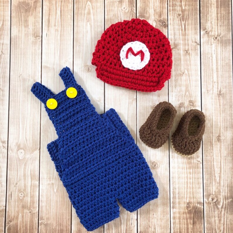 May include: A crocheted baby outfit in a Super Mario theme. The outfit includes a red hat with a white 'M' on it, blue overalls, and brown shoes.