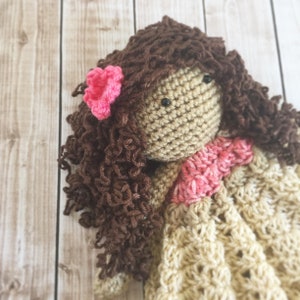 Pode incluir: Uma boneca de croch&ecirc; com cabelo castanho encaracolado, uma flor rosa e um vestido bege.