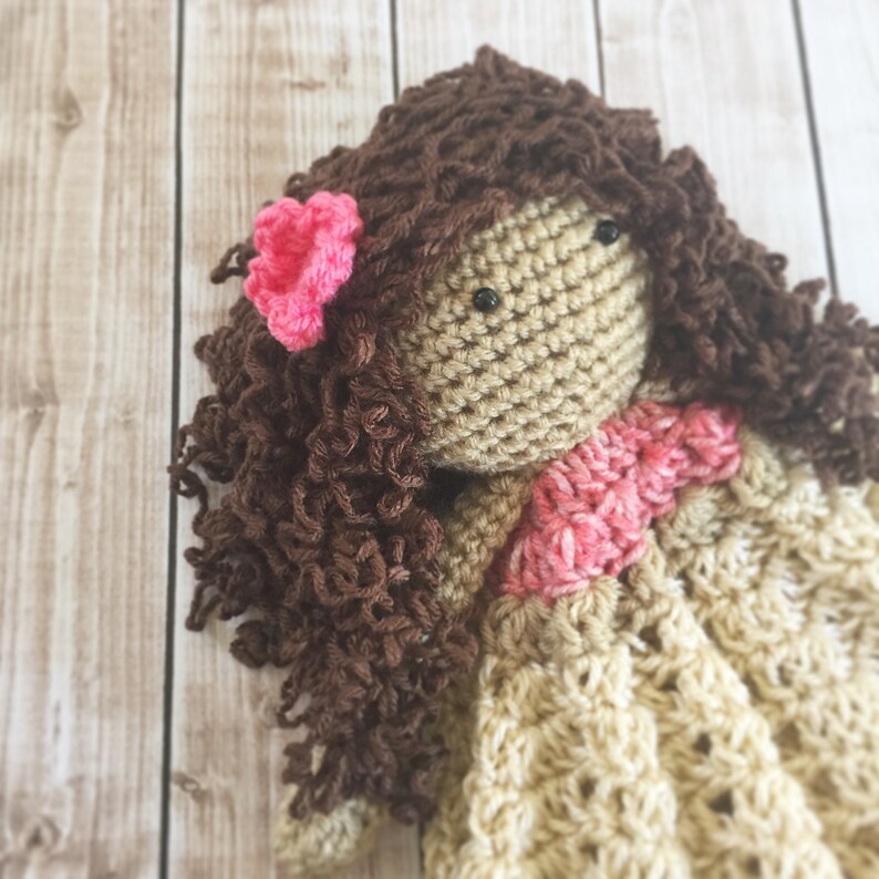 Pode incluir: Uma boneca de croch&ecirc; com cabelo castanho encaracolado, uma flor rosa e um vestido bege.