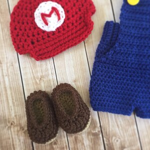Pu&ograve; includere: Un completo per neonato all'uncinetto in rosso, blu e marrone. Il completo comprende un cappello con una 'M' bianca, una salopette e scarpine.