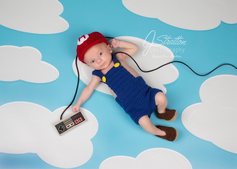 May include: A baby wearing a red and white crocheted hat and blue overalls is lying on a blue and white background with a white cord and a vintage video game controller.