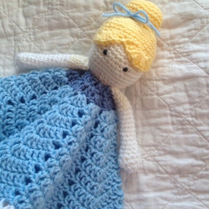 May include: A crocheted doll with a blue and white dress, yellow hair, and a blue bow. The doll is lying on a white quilted fabric.