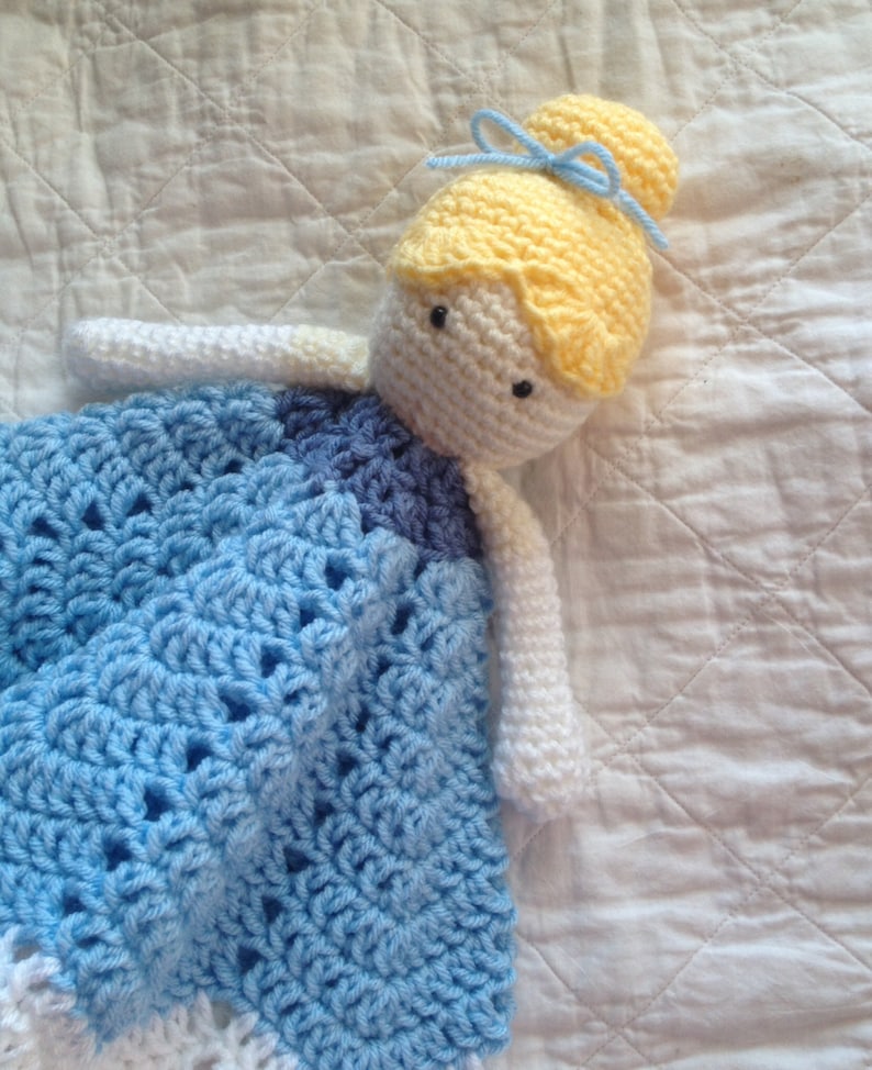 May include: A crocheted doll with a blue and white dress, yellow hair, and a blue bow. The doll is lying on a white quilted fabric.