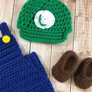 May include: A green crocheted hat with a white 'L' on the front, a blue crocheted overall with a yellow button, and two brown crocheted shoes.