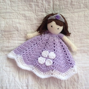May include: A crocheted doll wearing a purple dress with white flowers and a crown. The doll is lying on a white blanket.