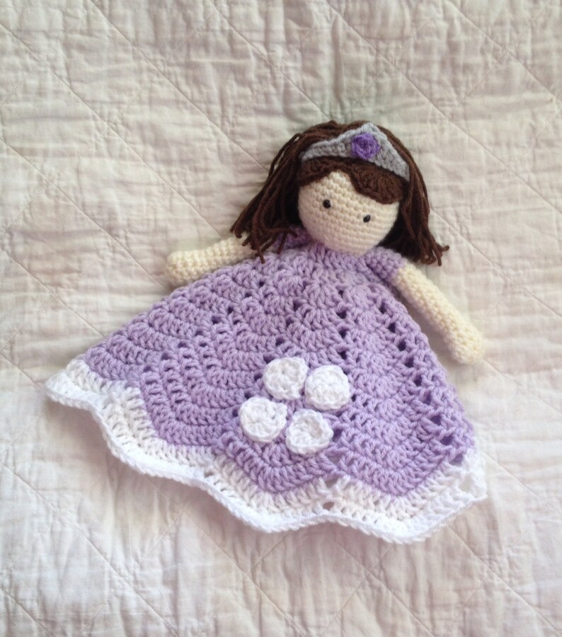 May include: A crocheted doll wearing a purple dress with white flowers and a crown. The doll is lying on a white blanket.