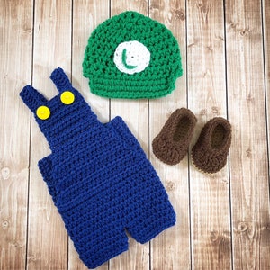 May include: A crocheted baby outfit in blue and green with a hat, overalls, and shoes. The hat has a green and white 'L' on it.