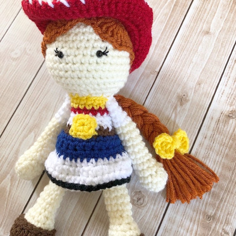 May include: Crocheted doll with a red and white cowboy hat, a white shirt with a yellow star, a blue skirt, and brown hair in a braid with yellow bows.